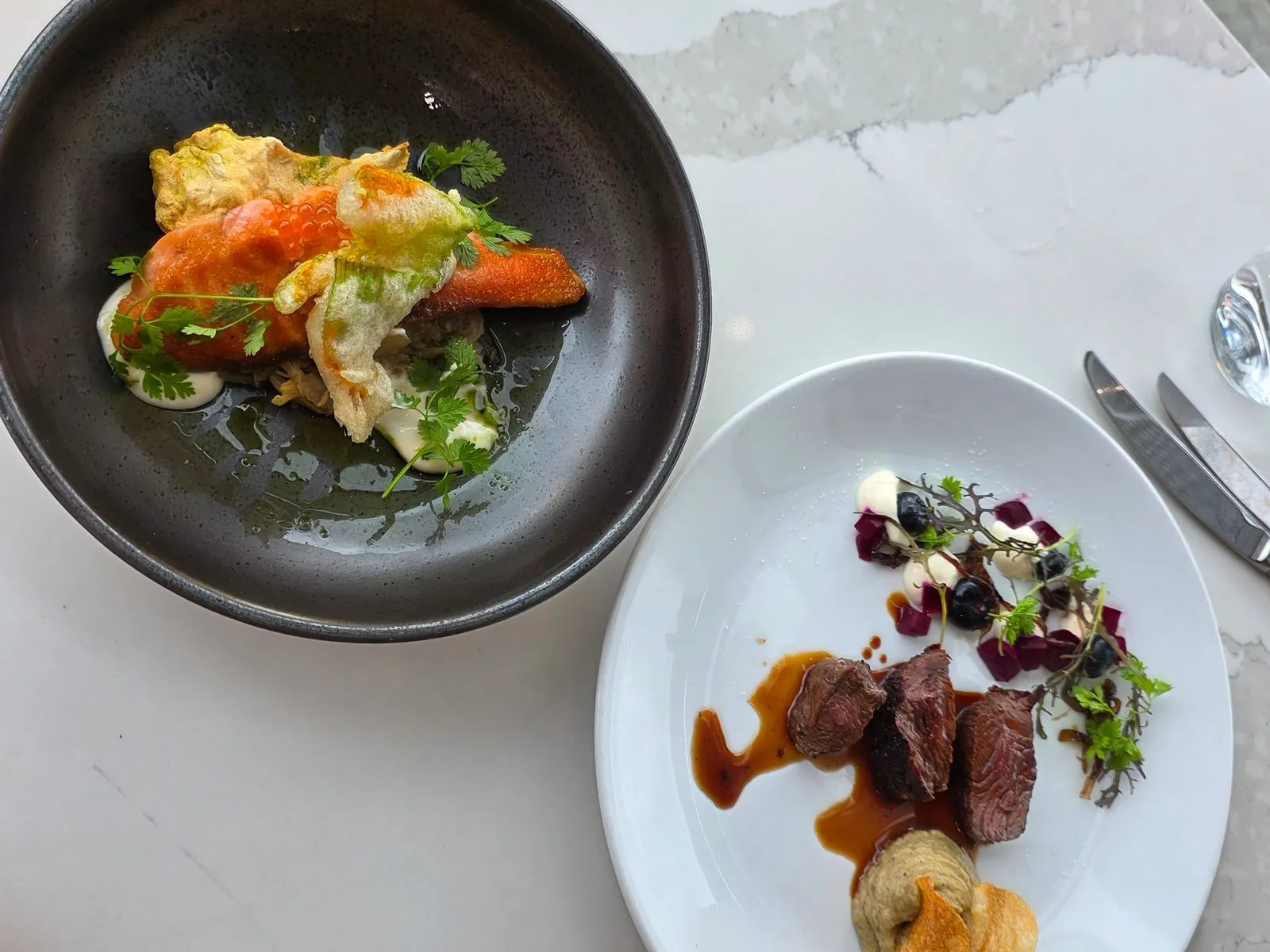 Two plated courses on a marble table, one on white china and one on dark ceramic